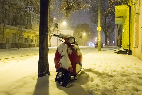Скутер посыпался снегом, стоя под деревом в свете ночных фонарей на фоне сильного снега и толстого слоя снега на земле. Ночь зимние улицы города фото города
