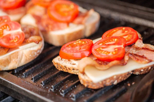 Grilling grilled cheese sandwich with bacon strips and fresh tomato on outdoor gas grill.