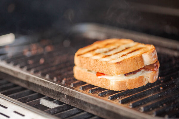 Grilling grilled cheese sandwich with bacon strips and fresh tomato on outdoor gas grill.