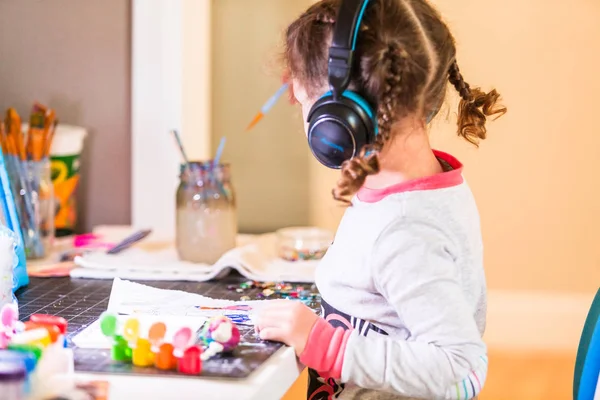 小女孩戴着耳机听音乐和工作在办公桌在家里的艺术项目 — 图库照片