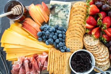 Aperatif olarak sunulan tahtanın üzerinde gurme peyniri, kraker ve meyve var..