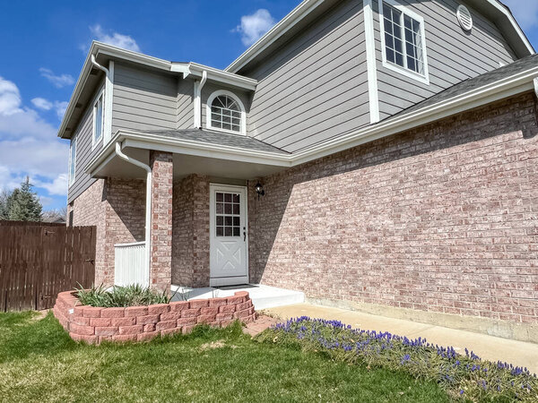 Двухэтажный кирпичный дом с белой отделкой и клумбами вдоль фундамента. Небо ярко-голубое с небольшими облаками и теплым солнечным светом.