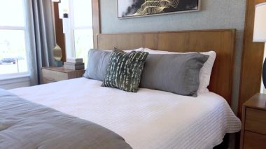 Kamera bir yatak odasının yan açısını yakalıyor. İçinde tahta bir yatak başlığı, beyaz yatak örtüsü ve yumuşak ışıklandırma var. Geniş pencere doğal ışığın boşluğu doldurmasına izin verir.