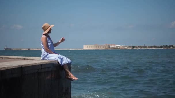 Mooie jongedame in een blauwe jurk zit op een houten pier op het meer — Stockvideo