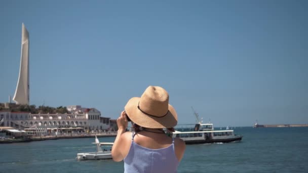 Vrouw met een hoed neemt foto's van de zee op haar mobiele telefoon op het strand — Stockvideo