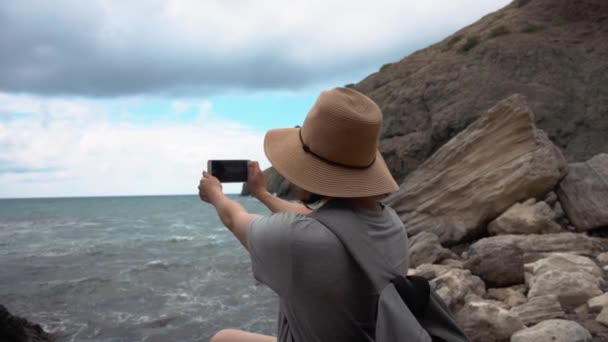 Vrouw met een hoed neemt foto's van de zee op haar mobiele telefoon op het strand — Stockvideo