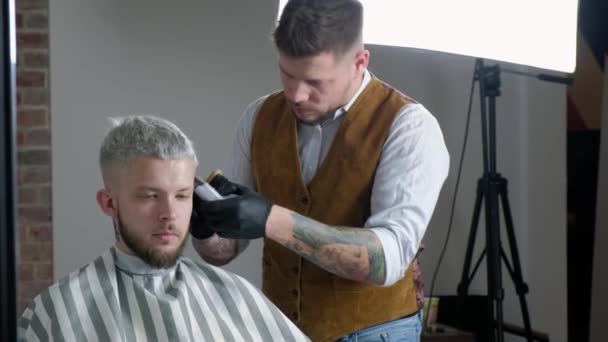 Att göra frisyr. Ung bearded man få frisyr av barber sittande i stol på frisersalong. — Stockvideo