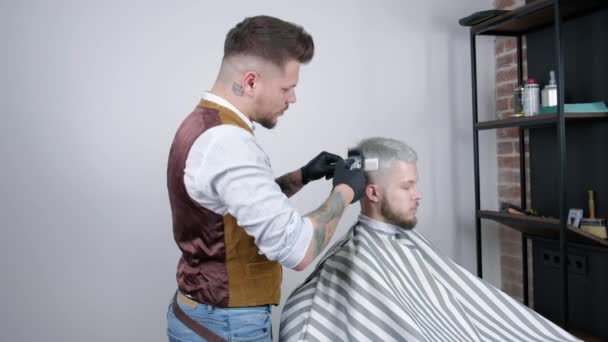 Att göra frisyr. Ung bearded man få frisyr av barber sittande i stol på frisersalong. — Stockvideo