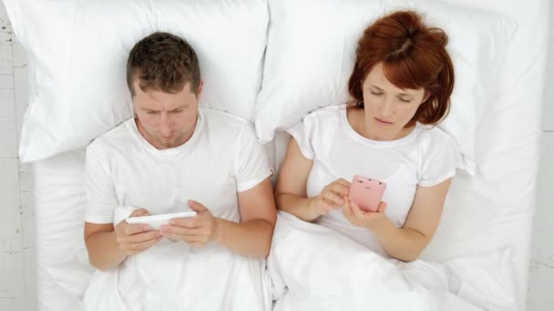 Par av makarna är i sängen och titta på Smartphones - kommunicerar i sociala nätverk och skriver meddelanden ligger i sängen före sänggåendet. — Stockvideo