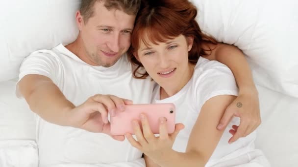 Ovanifrån på unga attraktiva paret liggande i sängen och ta bra selfies på din smartphone. — Stockvideo