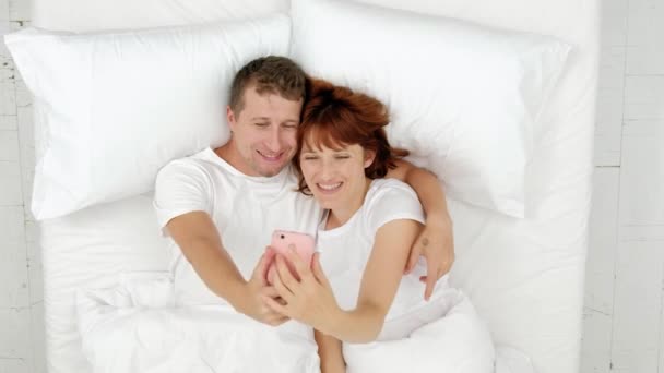 Vista superior de la joven pareja atractiva tumbada en la cama y tomando buenos selfies en el teléfono inteligente . — Vídeos de Stock