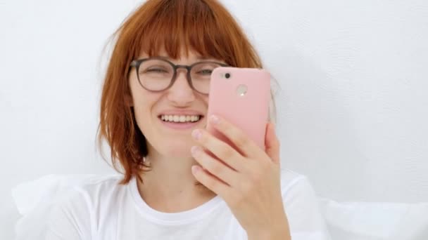 Portret van een meisje met een telefoon, uw favoriete deuntje in een slimme telefoon smiling zoeken, beluisteren door de koptelefoon, geïsoleerd op een witte achtergrond. — Stockvideo