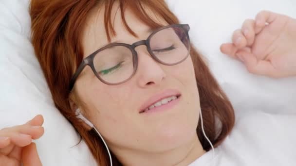 Ritratto dall'alto di bella, attraente, bruna, affascinante, ragazza allegra che si diverte con lo smartphone, auricolare sulla testa, ascoltando la sua playlist preferita, scegliendo, cercando la canzone, sdraiato sul retro . — Video Stock