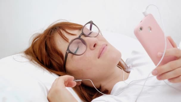 Top view portrait of pretty, attractive, brunette, charming, cheerful girl having fun using smart phone, headset on head, listening her favorite playlist, choosing, searching for song, lying on back. — Stock Video