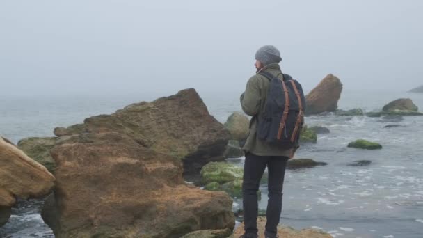 Widok z tyłu jednego samotnego dorosłego faceta spędzać czas przez chodzenie i rozejrzeć się na zdjęciach piękno dzikiej nadmorskiej okolicy. Zielone wybrzeże nabrzeża, zimna wietrzna pogoda, chmury zamykające horyzont — Wideo stockowe