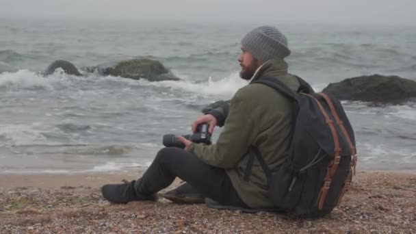Hombre barbudo tomando fotos con cámara profesional en frente del océano — Vídeo de stock