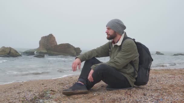 Young bearded man sits alone on beach in the evening and throws pebbles into the water — 비디오