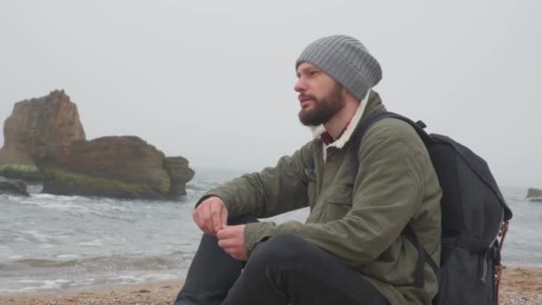 Young bearded man sits alone on beach in the evening and throws pebbles into the water — 비디오