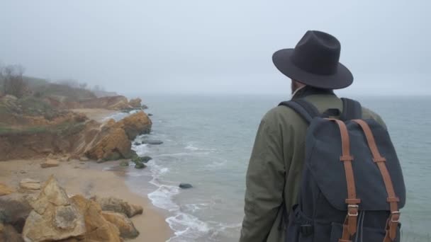 Backview of a fashionable guy in a hat and jacket taking pictures of beautiful natural scenes using a seaside mobile phone. Wild cold day — Stock Video