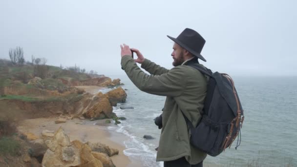 Backview τύπος με ένα καπέλο και μπουφάν λήψη φωτογραφίες από όμορφες φυσικές σκηνές χρησιμοποιώντας ένα παραθαλάσσιο κινητό τηλέφωνο. Άγρια κρύα μέρα — Αρχείο Βίντεο