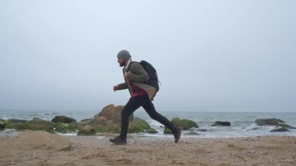 Bebaarde hipster reiziger met een rugzak wandelingen langs het strand — Stockvideo