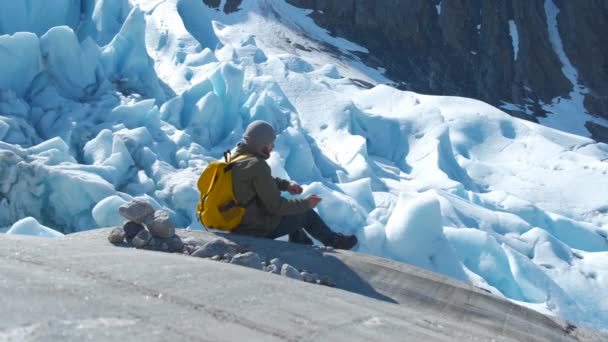 Turista con uno zaino giallo guarda un bellissimo ghiacciaio. getta pietre — Video Stock