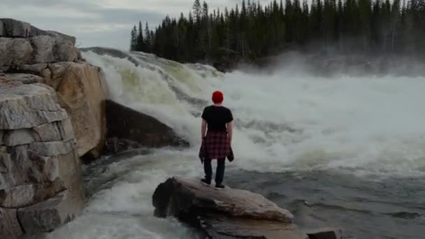 Travel man staande in de voorkant van de waterval in Noorwegen bos in de vroege ochtend. Gekleed in een rode hoed. — Stockvideo