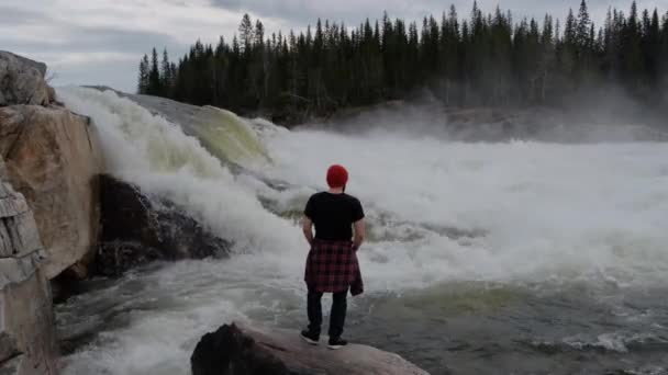 Resa man stående framför vattenfall i Norge skog på tidig morgon. Klädd i en röd hatt. — Stockvideo