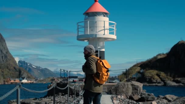 Travel man met een gele rugzak gaat op een pittoresk heuvelachtig terrein naar de vuurtoren in de buurt van de Oceaan — Stockvideo