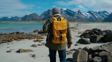 Büyük dağlar ve arka planda kırma dalgaları ile sarı sırt çantası taşıyan uzak Kum Plajı üzerinde yürüyüş yeşil ceket giyen Hiker, Lofoten Adaları Norveç