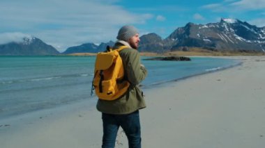 Büyük dağlar ve arka planda kırma dalgaları ile sarı sırt çantası taşıyan uzak Kum Plajı üzerinde yürüyüş yeşil ceket giyen Hiker, Lofoten Adaları Norveç