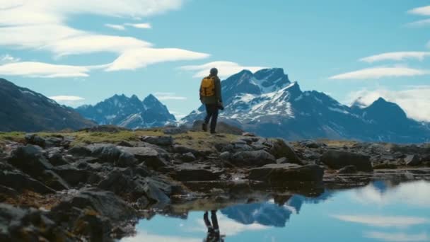 Young bearded man in a hat with a backpack comes down from the mountains on a background of snow capped mountains — Stock Video