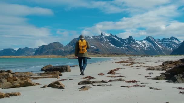 Пешеходный турист в зеленой куртке, идущий по удаленному песчаному пляжу с желтым рюкзаком с большими горами и ломающимися волнами на заднем плане, Норвегия — стоковое видео