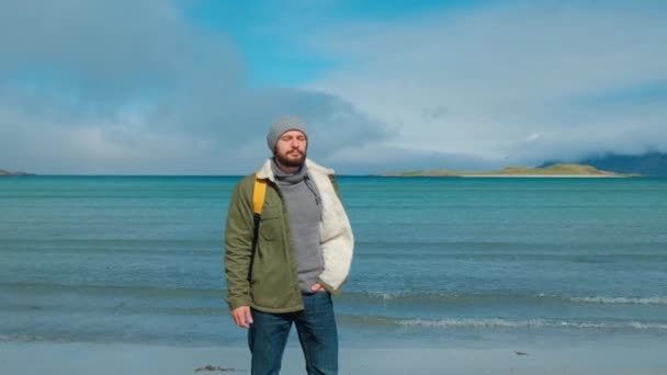 Voyageur vêtu d'une veste verte et d'un sac à dos jaune se dresse sur la plage et sur fond de sommets de haute montagne, inspiré par le paysage épique des îles Lofoten, en Norvège — Video