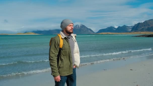 Voyageur vêtu d'une veste verte et d'un sac à dos jaune se dresse sur la plage et sur fond de sommets de haute montagne, inspiré par le paysage épique des îles Lofoten, en Norvège — Video