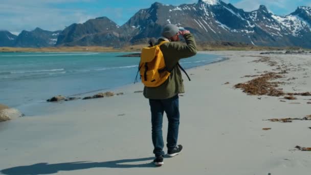 Caminhante vestindo jaqueta verde andando na praia de areia remota carregando mochila amarela com grandes montanhas e quebrando ondas no fundo, Lofoten Islands Norway — Vídeo de Stock