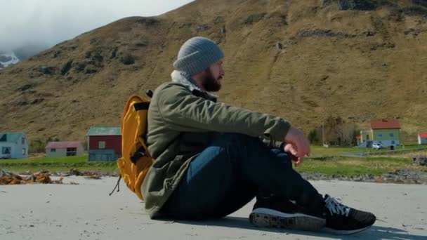 Homme barbu assis sur une plage de sable avec sac à dos jaune, hipster bénéficie de la vue sur la mer bleue et regarde au loin, les vagues battent sur le rivage — Video