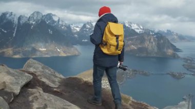 Yavaş hareket içinde gün batımında bir dağın zirvesinde yürüyen adam. Ceket, kırmızı şapka ve sarı backpac takıyor. Norveç Lofoten