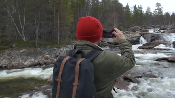 Resenären i en röd hatt med en ryggsäck full rygg stannade för att ta ett foto av en Bergs flod. konceptet resor och fotografi. — Stockvideo