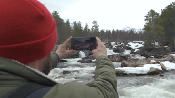 Voyageur en chapeau rouge avec un sac à dos plein à l'arrière arrêté pour prendre une photo d'une rivière de montagne. concept de voyage et de photographie . — Video