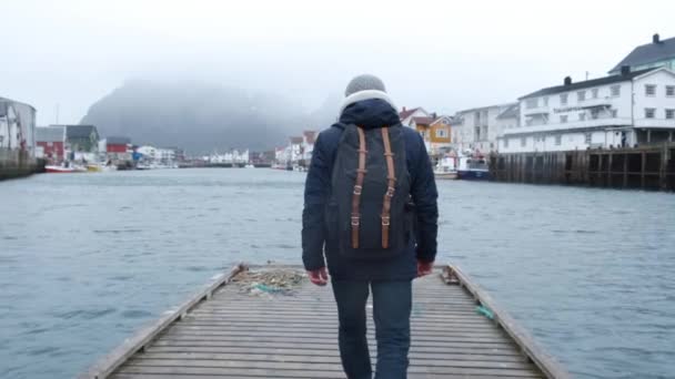 Viajante vai em um cais de madeira. vestido com uma jaqueta e chapéu de malha e mochila. vista para trás. Noruega Reine — Vídeo de Stock