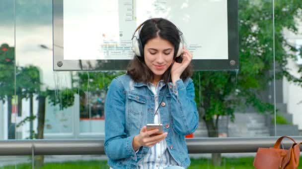 Giovane donna in cuffia godendo la musica, in attesa dei mezzi pubblici mentre si siede alla moderna stazione del tram all'aperto — Video Stock