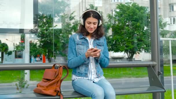 Mujer joven en auriculares disfrutando de la música, esperando el transporte público mientras se sienta en la moderna estación de tranvía al aire libre — Vídeos de Stock