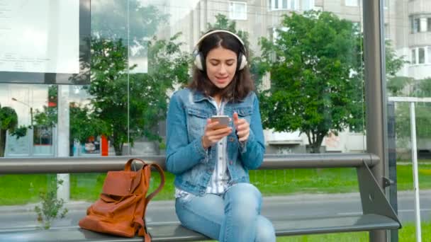 Junge Frau mit Kopfhörern genießt Musik und wartet auf die öffentlichen Verkehrsmittel, während sie an der modernen Straßenbahnhaltestelle im Freien sitzt — Stockvideo