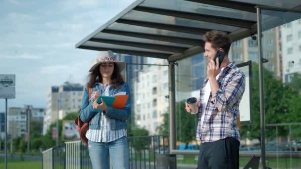 年轻人和女孩。 站在巴士站上。 太阳在照耀着。 男人用他的手机 在等他的电车 城市生活方式. — 图库视频影像