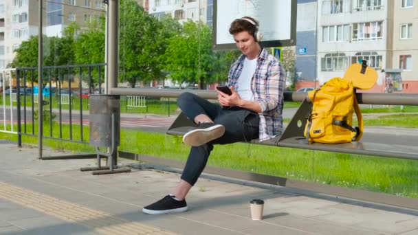 Radosny młody człowiek czeka na transport na przystanku autobusowym. mężczyzna siedzi na ławce. Facet za pomocą smartphone słucha muzyki — Wideo stockowe