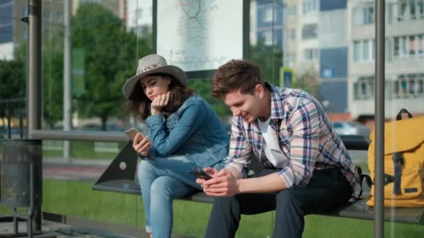 Mann und Frau sitzen zusammen am Bahnhof. sie halten moderne Geräte in der Hand und unterhalten mit Internet, während sie auf die Abfahrt warten. — Stockvideo