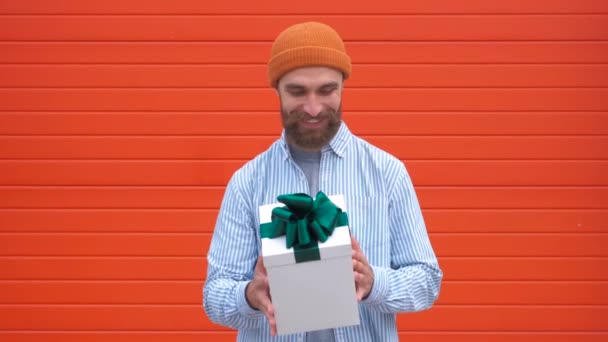 Joven hipster sorprendido con bigote y barba en sorpresa sosteniendo caja blanca con regalos sobre un fondo rojo, El concepto de regalos y sorpresas para las vacaciones — Vídeo de stock