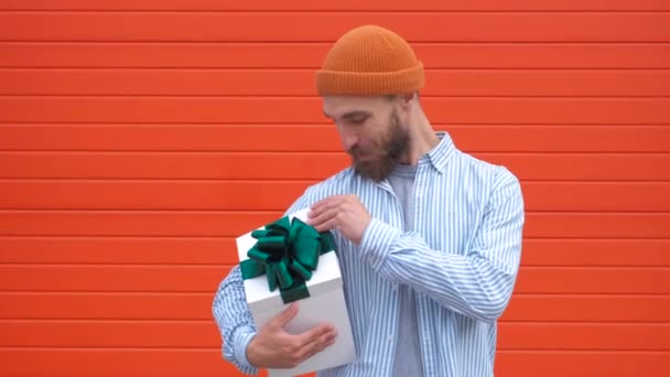 Guapo joven hipster sosteniendo caja de regalo sobre un fondo rojo. Concepto de vacaciones y cumpleaños — Vídeo de stock