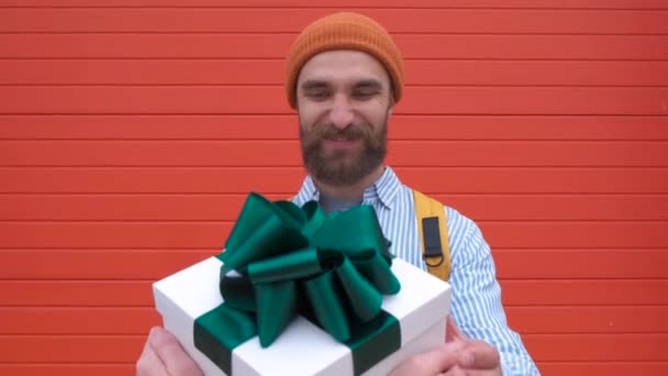 Porträt eines aufgeregten bärtigen Mannes, der sich freut, weiße Geschenkschachtel mit grüner Schleife aus weiblicher Hand über rote Wand zu bekommen — Stockvideo